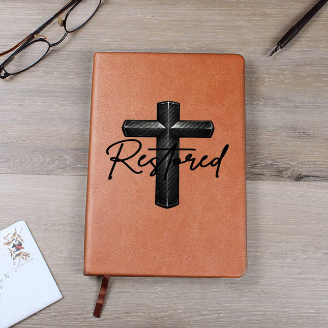 Tan vegan leather hardcover journal with a carbon-fiber black cross graphic and the word "Restored" in black script on the cover, displayed on a light wood surface with glasses and a pen nearby.