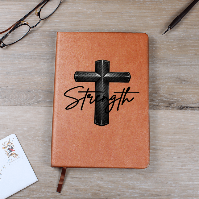 Tan vegan leather hardcover journal with a carbon-fiber black cross graphic and the word "Strength" in black script on the cover, displayed on a light wood surface with glasses and a pen nearby.