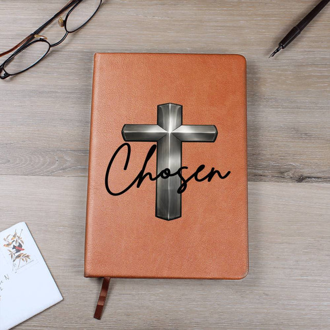 Tan vegan leather hardcover journal with a silver chrome 3D cross graphic and the word "Chosen" in black script on the cover, displayed on a light wood surface with glasses and a pen nearby.