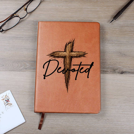 Tan vegan leather hardcover journal with a rustic wooden cross graphic and the word "Devoted" in black script on the cover, displayed on a light wood surface with glasses and a pen nearby.