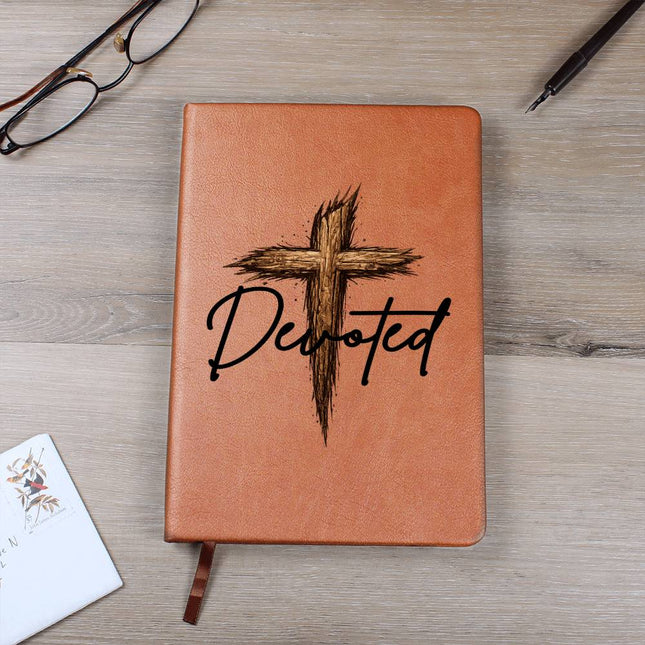 Tan vegan leather hardcover journal with a rustic wooden cross graphic and the word "Devoted" in black script on the cover, displayed on a light wood surface with glasses and a pen nearby.