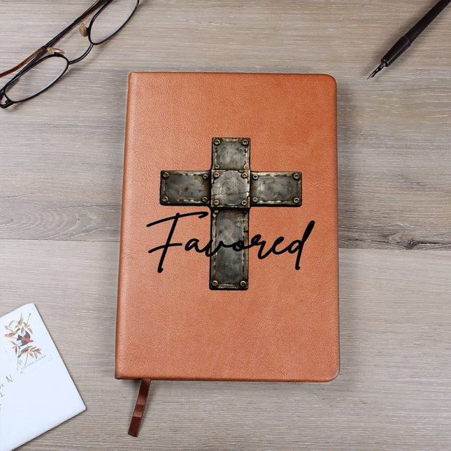 Tan vegan leather hardcover journal with a vintage stone-tile riveted cross graphic and the word "Favored" in black script on the cover, displayed on a light wood surface with glasses and a pen nearby.