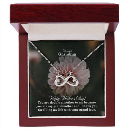 Heart-shaped infinity endless love necklace in white gold finish held in hands, displayed in a gift box with a Dearest Grandma Happy Mother's Day message card and soft pink dahlia flower background.