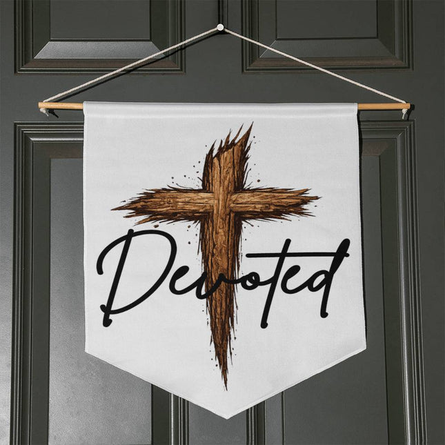 White pennant-shaped wall hanging with a rustic wooden cross graphic and the word "Devoted" in black script, hung with a wooden dowel and twine on a dark door.