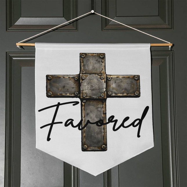 White pennant-shaped wall hanging with a vintage stone-tile riveted cross graphic and the word "Favored" in black script, hung with a wooden dowel and twine on a dark door.