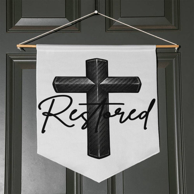 White pennant-shaped wall hanging with a carbon-fiber black cross graphic and the word "Restored" in black script, hung with a wooden dowel and twine on a dark door.