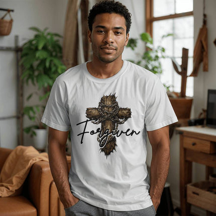 Man wearing a white unisex t-shirt featuring a rugged wooden cross wrapped with a crown of thorns and chains with the word "Forgiven" in black script on the front, styled in a modern home setting.