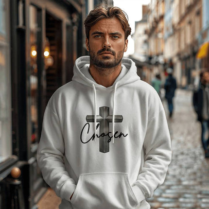 Man wearing a white unisex pullover hoodie featuring a silver chrome 3D cross graphic with the word "Chosen" in black script on the chest, styled on a cobblestone city street.