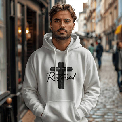 Man wearing a white unisex pullover hoodie featuring a carbon-fiber black cross graphic with the word "Restored" in black script on the chest, styled on a cobblestone city street.