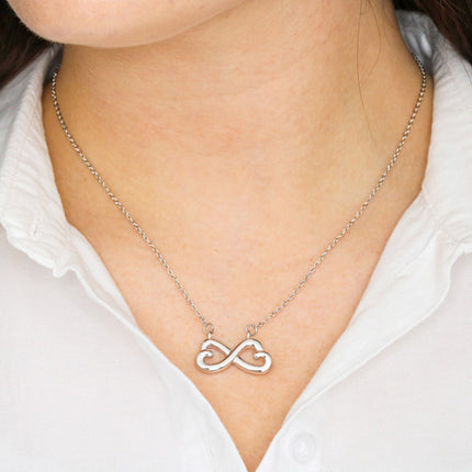 Heart-shaped infinity endless love necklace in white gold finish held in hands, displayed in a gift box with a Dearest Grandma Happy Mother's Day message card and soft pink dahlia flower background.