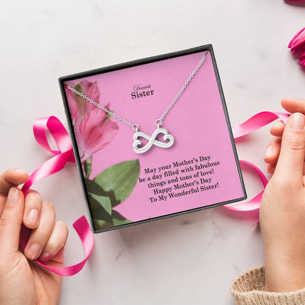 Heart-shaped infinity endless love necklace in white gold finish held in hands, displayed in a gift box with a Dearest Sister Happy Mother's Day message card on a pink floral background.