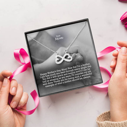 Heart-shaped infinity endless love necklace in white gold finish held in hands, displayed in a gift box with a Dear Mom Happy Mother's Day message card and black and white mother and baby photo background.
