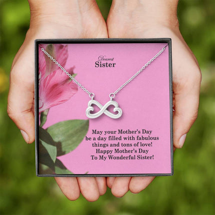 Heart-shaped infinity endless love necklace in white gold finish held in hands, displayed in a gift box with a Dearest Sister Happy Mother's Day message card on a pink floral background.