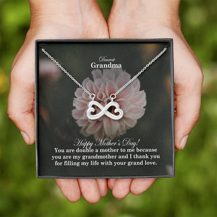Heart-shaped infinity endless love necklace in white gold finish held in hands, displayed in a gift box with a Dearest Grandma Happy Mother's Day message card and soft pink dahlia flower background.