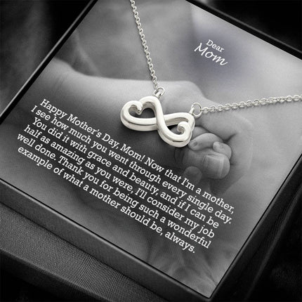 Heart-shaped infinity endless love necklace in white gold finish held in hands, displayed in a gift box with a Dear Mom Happy Mother's Day message card and black and white mother and baby photo background.