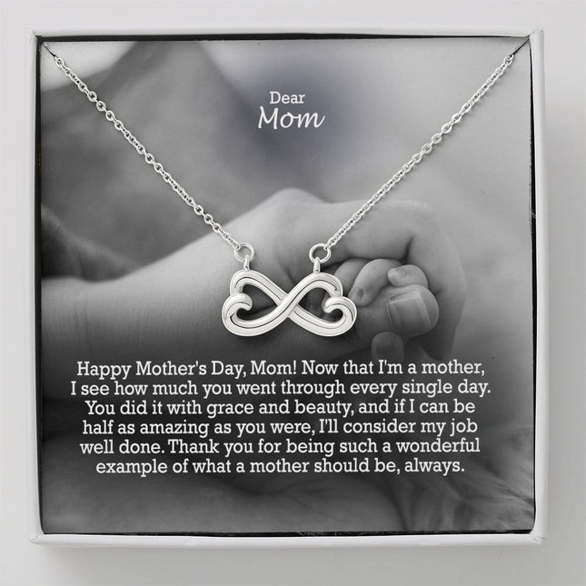 Heart-shaped infinity endless love necklace in white gold finish held in hands, displayed in a gift box with a Dear Mom Happy Mother's Day message card and black and white mother and baby photo background.