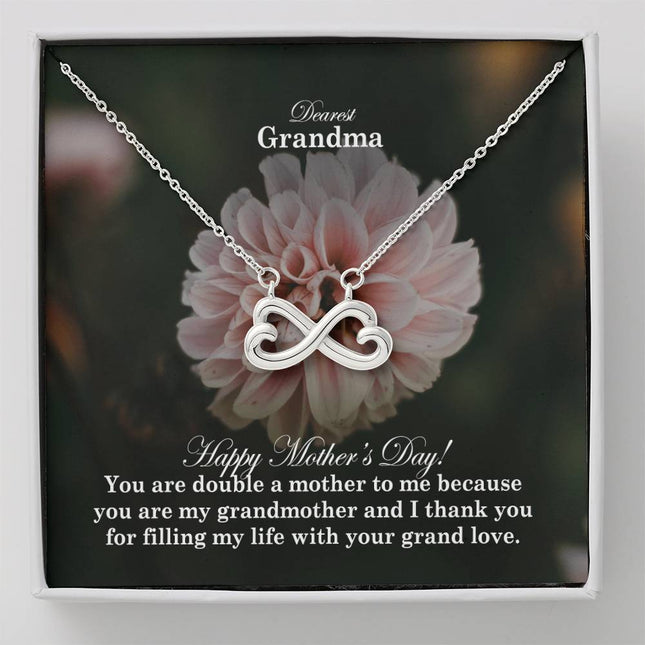 Heart-shaped infinity endless love necklace in white gold finish held in hands, displayed in a gift box with a Dearest Grandma Happy Mother's Day message card and soft pink dahlia flower background.