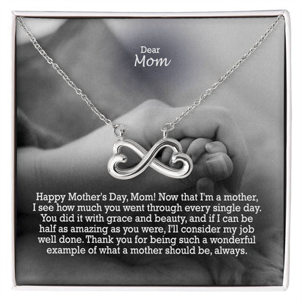 Heart-shaped infinity endless love necklace in white gold finish held in hands, displayed in a gift box with a Dear Mom Happy Mother's Day message card and black and white mother and baby photo background.