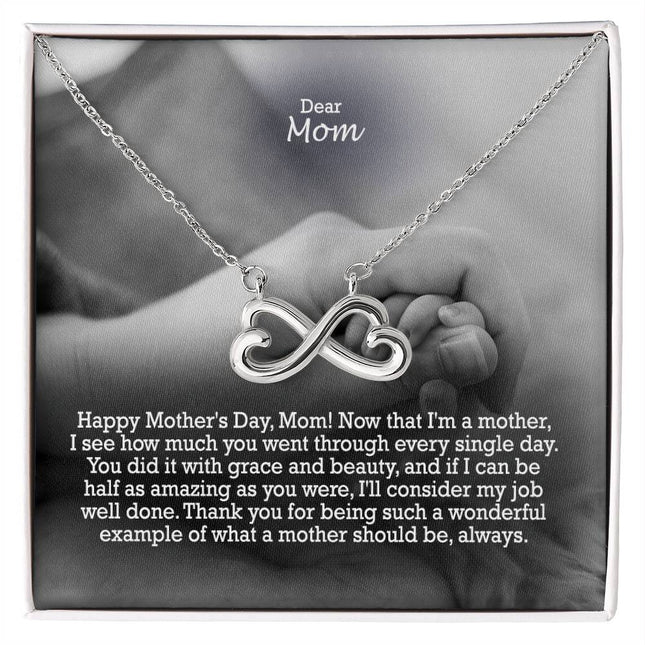 Heart-shaped infinity endless love necklace in white gold finish held in hands, displayed in a gift box with a Dear Mom Happy Mother's Day message card and black and white mother and baby photo background.