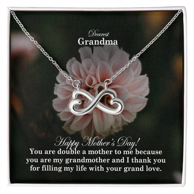 Heart-shaped infinity endless love necklace in white gold finish held in hands, displayed in a gift box with a Dearest Grandma Happy Mother's Day message card and soft pink dahlia flower background.