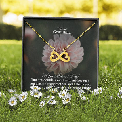 Heart-shaped infinity endless love necklace in white gold finish held in hands, displayed in a gift box with a Dearest Grandma Happy Mother's Day message card and soft pink dahlia flower background.