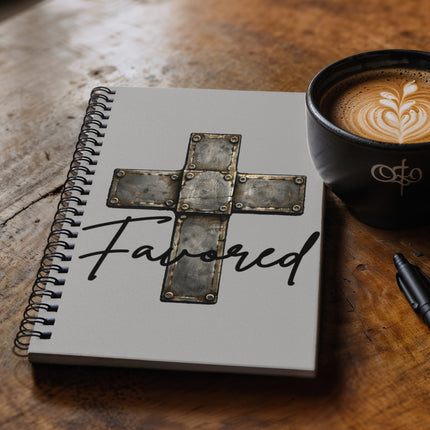 Light gray spiral notebook with a vintage stone-tile riveted cross graphic and the word "Favored" in black script on the cover, displayed on a rustic wood surface beside a black coffee cup.