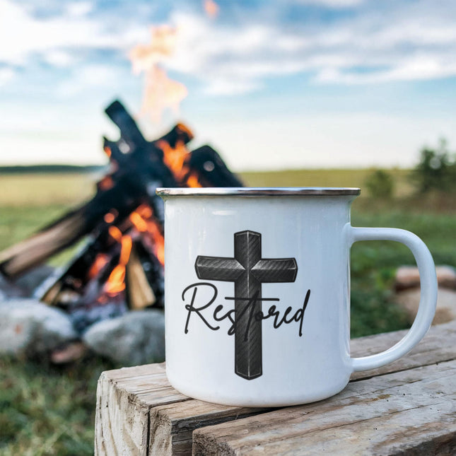 White enamel camping mug with a carbon-fiber black cross graphic and the word "Restored" in black script, sitting on a rustic wood stump with a campfire blazing in the background.