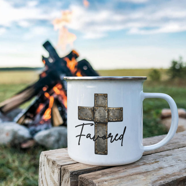 White enamel camping mug with a vintage stone-tile riveted cross graphic and the word "Favored" in black script, sitting on a rustic wood stump with a campfire blazing in the background.