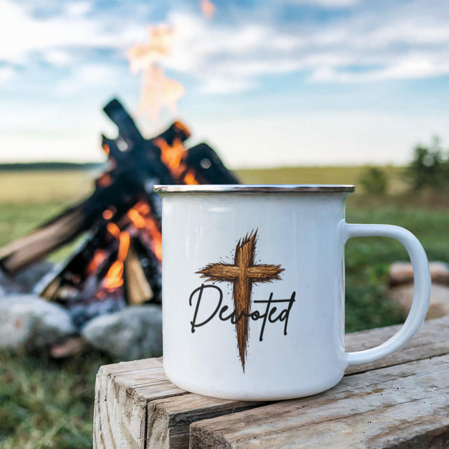 White enamel camping mug with a rustic wooden cross graphic and the word "Devoted" in black script, sitting on a rustic wood stump with a campfire blazing in the background.