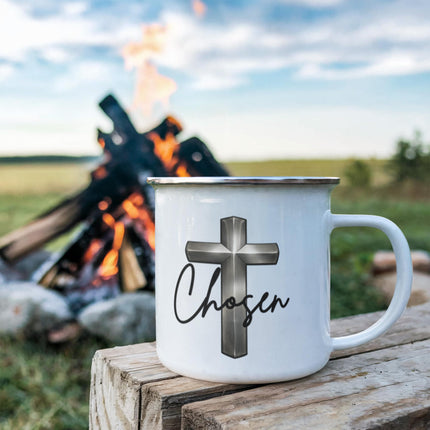 White enamel camping mug with a silver chrome cross graphic and the word "Chosen" in black script, sitting on a rustic wood stump with a campfire blazing in the background.