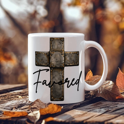 White ceramic coffee mug with a vintage stone-tile riveted cross graphic and the word "Favored" in black script, displayed on a rustic wooden surface surrounded by autumn leaves.
