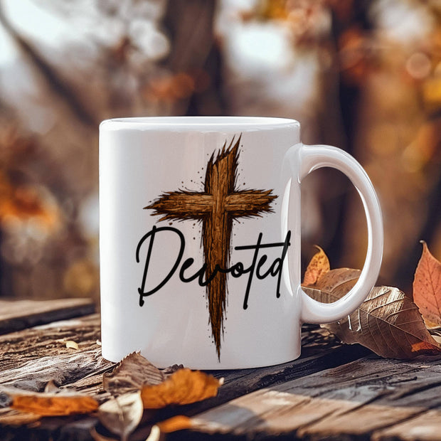 White ceramic coffee mug with a rustic wooden cross graphic and the word "Devoted" in black script, displayed on a rustic wooden surface surrounded by autumn leaves.