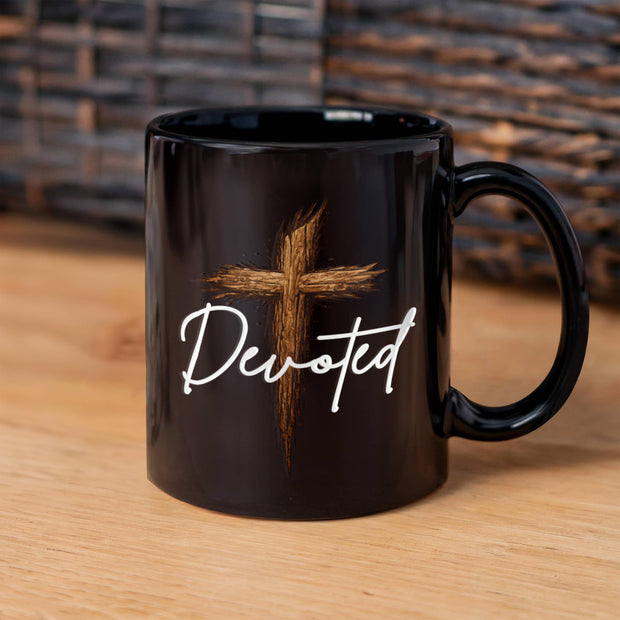 Glossy black ceramic coffee mug with a rustic wooden cross graphic and the word "Devoted" in white script, displayed on a light wood surface with a wicker backdrop.